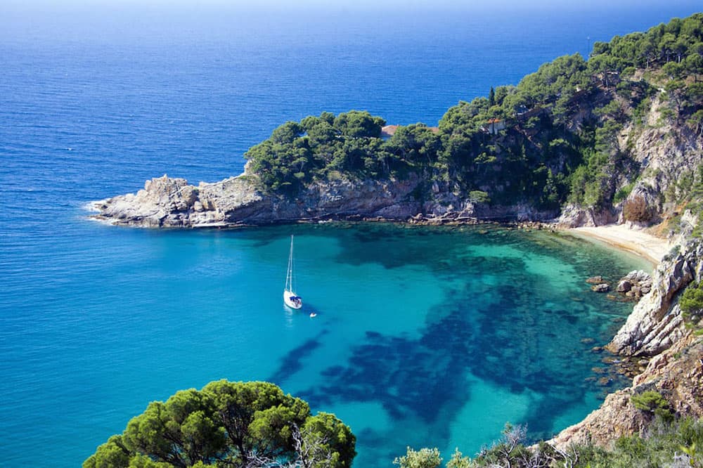A serene coastal scene with a single Hanse sailboat on clear turquoise waters near a tree-covered cliff at turkish riviera