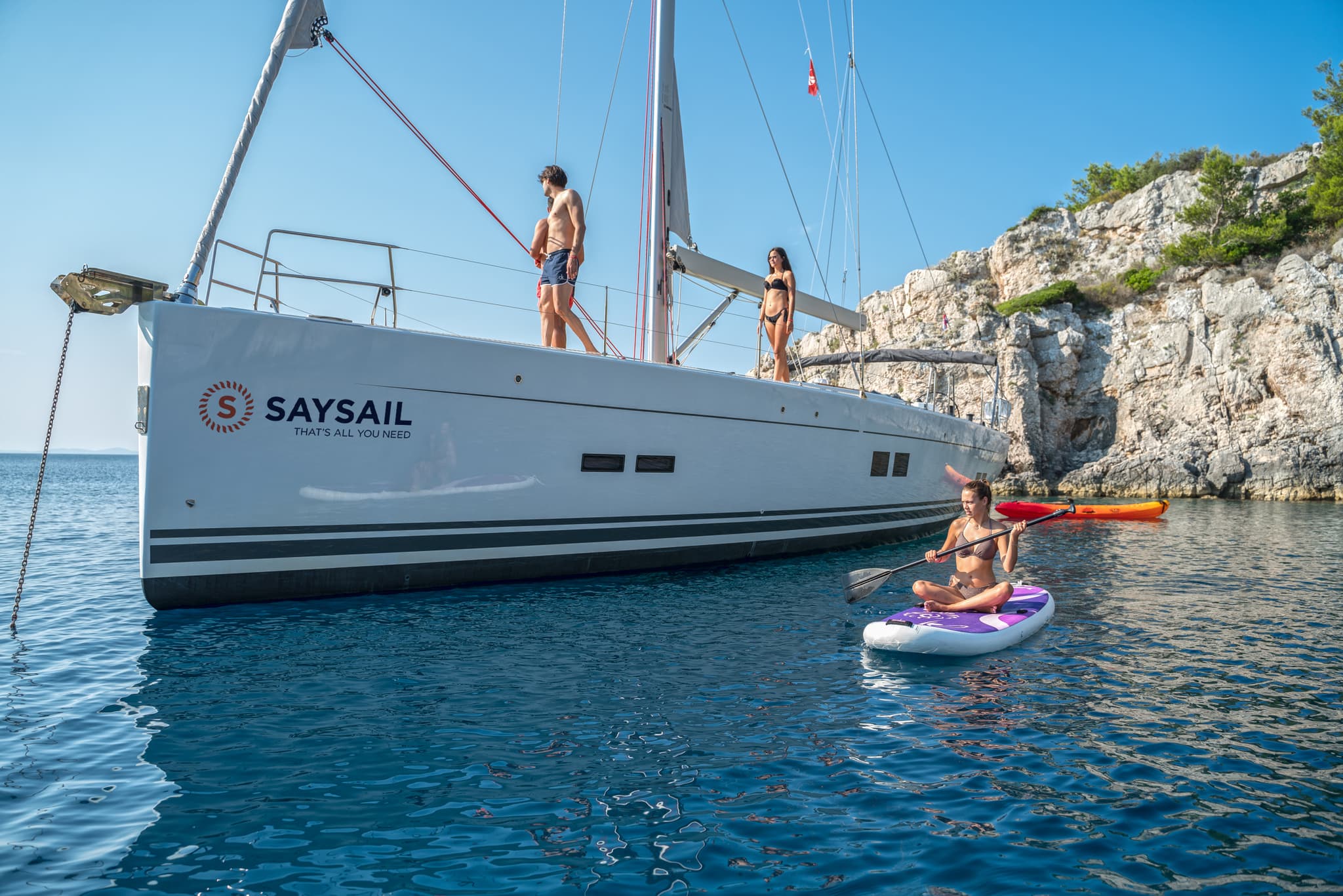 People enjoying water activities near a sailingboat anchored by a rocky coast