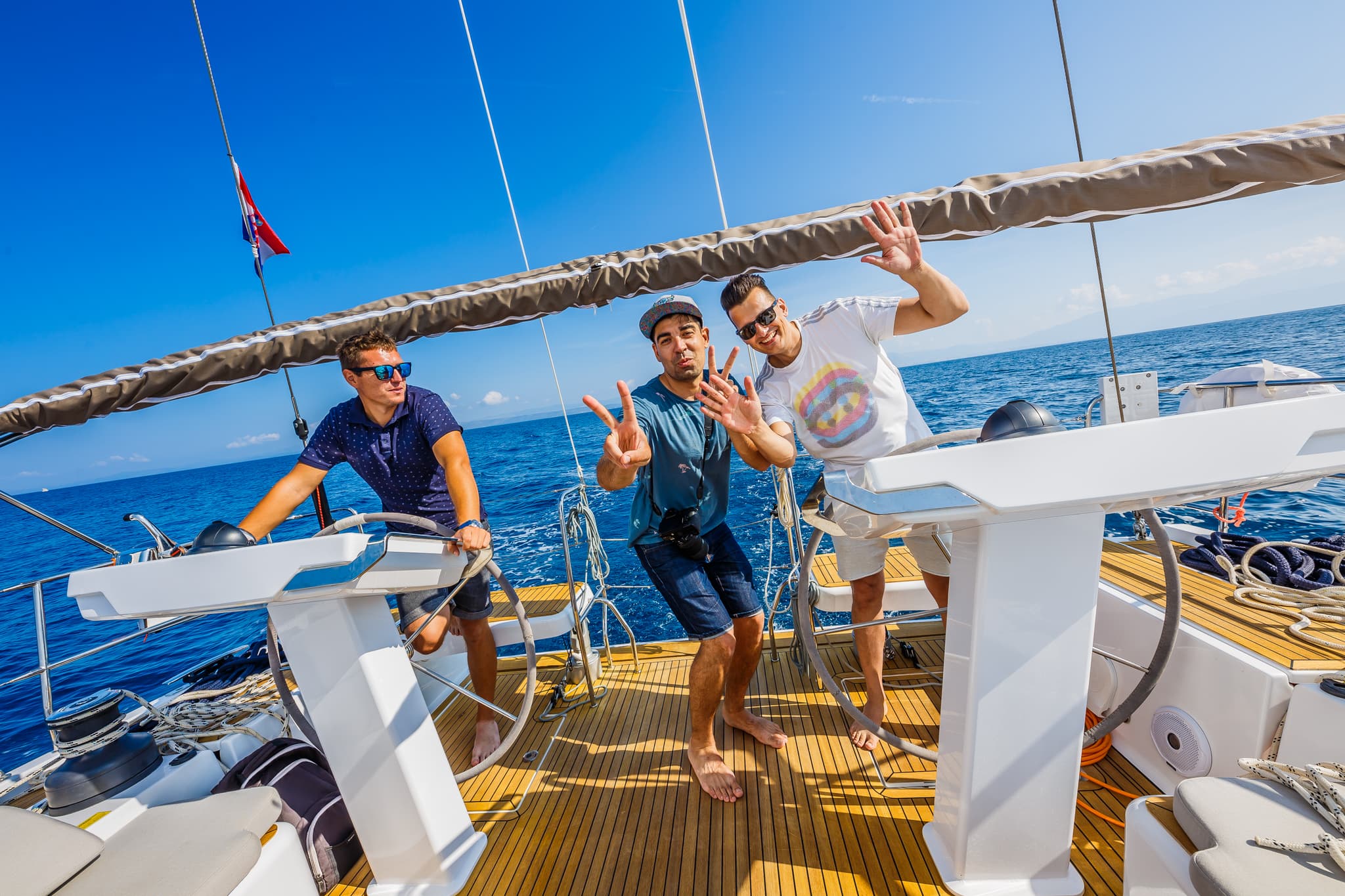 Three men on a Hanse sailing yacht deck, waving under clear blue skies