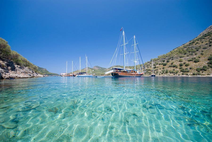Crystal clear waters with a traditional sailboat anchored near a rocky coastline under a blue sky